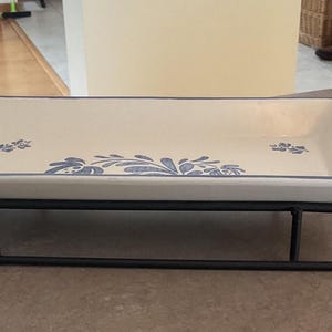 May include: A rectangular serving dish with a cream-colored interior and blue floral designs, set on a black metal sleigh-style stand. The dish has a blue border and curved handles. A decorative piece for serving food.