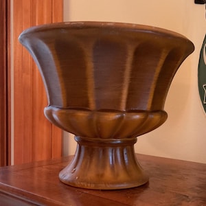 May include: A large, brown, decorative urn with a fluted design. The urn has a wide, flared top and a pedestal base. The color is a warm, earthy tone, and the surface appears to have a slightly aged finish. The urn is likely made of ceramic or a similar material.