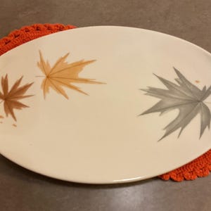 May include: An oval, off-white ceramic platter with three painted maple leaves in brown, orange, and gray. The platter rests on a round, orange crocheted doily. A fall-themed decorative piece.