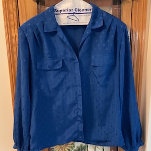 May include: A long-sleeved button-down shirt in a vibrant royal blue color featuring a subtle tonal square pattern throughout the fabric. The shirt includes a classic collar, two front chest pockets with flaps, and long sleeves with cuffs.
