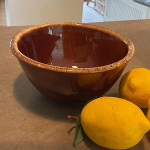 May include: A deep, glossy brown ceramic bowl with a speckled, lighter tan rim. The bowl has a smooth, reflective interior and exterior surface. Next to the bowl are two bright yellow, textured decorative lemons with a small green leaf attached.