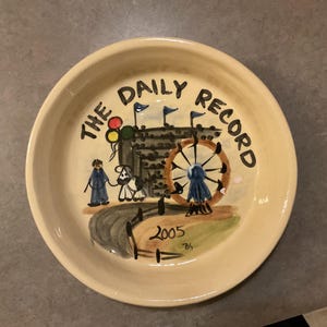 May include: Cream-colored ceramic plate with a hand-painted design. The plate features the text "THE DAILY RECORD" in black, along with a Ferris wheel, figures, and the year 2005. The rim of the plate is slightly raised.