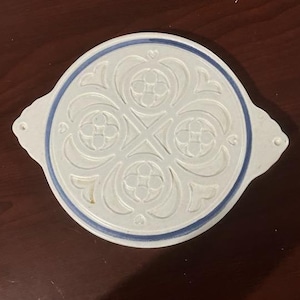 May include: A white ceramic trivet with a blue rim and an intricate embossed design. The trivet has two handles on the sides.