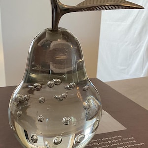 May include: A clear glass pear-shaped decorative object with embedded bubbles. It has a dark metal stem and leaf detail. The pear is sitting on a brown book.