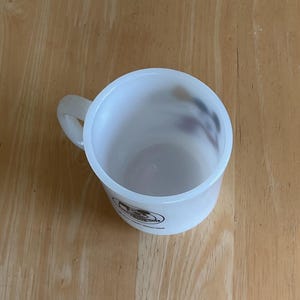 Puede incluir: Una taza de pl&aacute;stico blanco con asa, vista desde arriba. La taza tiene forma cil&iacute;ndrica y un aspecto ligeramente transl&uacute;cido. Un logotipo es visible en el lateral. La taza est&aacute; sobre una superficie de madera.