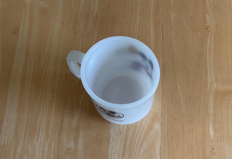 Puede incluir: Una taza de pl&aacute;stico blanco con asa, vista desde arriba. La taza tiene forma cil&iacute;ndrica y un aspecto ligeramente transl&uacute;cido. Un logotipo es visible en el lateral. La taza est&aacute; sobre una superficie de madera.