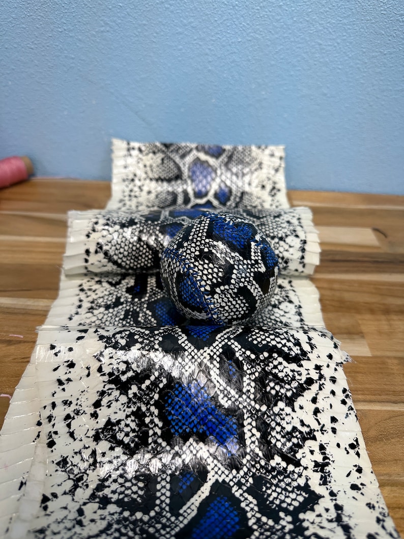 以下が含まれることがあります： 白、黒、青色のヘビ革模様の手作りアイテムのコレクション。ボールと折りたたまれた素材が含まれており、すべて木製の表面に表示されています。ヘビ革の模様は詳細で人目を引きます。