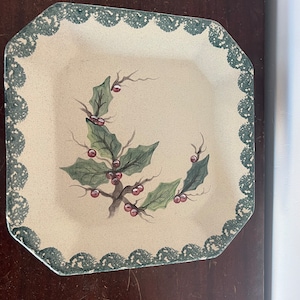 Może przedstawiać: Kwadratowy ceramiczny talerz z kremowym środkiem i zieloną, falistą obwódką. Talerz zdobi ręcznie malowany wzór ostrokrzewu z zielonymi liśćmi i czerwonymi jagodami, idealny na święta.