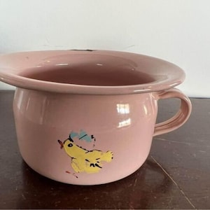 Op de afbeelding: Een roze emaille po met een handvat en een brede rand. Aan de zijkant is een gele kuiken met blauwe en rode accenten geschilderd. De pot is waarschijnlijk vintage en heeft een glanzende afwerking.