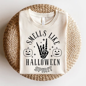 May include: A white t-shirt with a black graphic design that says "Smells Like Halloween Spirit". The graphic includes a skeleton hand making a rock and roll sign, two jack-o'-lantern pumpkins, and stars.