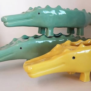 Puede incluir: Tres macetas de cerámica con forma de cocodrilo, una verde y dos verde y amarilla. Los cocodrilos verdes están apilados uno encima del otro, con el cocodrilo amarillo en frente.