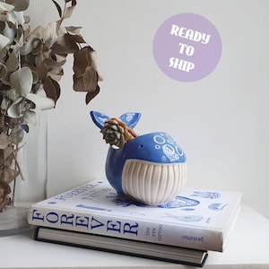 May include: A blue and white ceramic whale-shaped planter featuring a ribbed belly and a small succulent plant inside. The whale has a decorative bubble pattern on its head and is positioned on top of a white book titled Forever The New Tattoo.