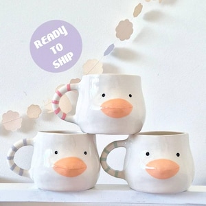 Puede incluir: Tres tazas de cerámica blanca con forma de pato, cada una con un pico naranja prominente, dos pequeños ojos negros y un asa con rayas horizontales en rosa, azul claro o verde. Se ve un círculo morado con el texto READY TO SHIP.