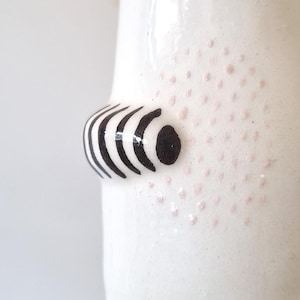 May include: A white ceramic vessel featuring a protruding, rounded element decorated with black horizontal stripes. The surface of the white ceramic is further embellished with a subtle pattern of small, light pink dots surrounding the striped protrusion.