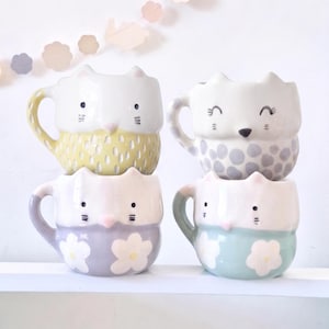Puede incluir: Cuatro tazas apiladas de cerámica con forma de gato. Cada taza es blanca con un diseño de cara de gato y una base de color. Las bases son amarillas con un patrón de puntos, grises con lunares, lavanda con flores y azul claro con flores.