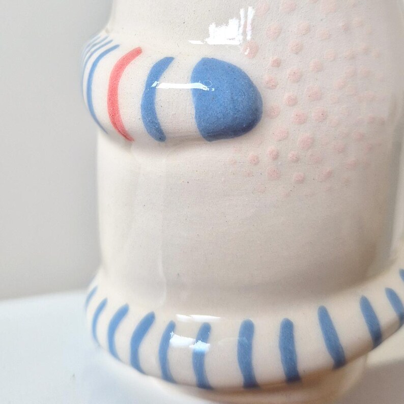 May include: A white ceramic vessel featuring a raised, rounded handle with blue and coral-colored stripes. The surface is decorated with a pattern of small, light pink dots and a base rim adorned with vertical blue stripes around the lower edge.