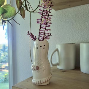 May include: A white ceramic cat-shaped bud vase with a hand-painted face, whiskers, and a striped tail. The vase features a textured belly and holds two tall, slender purple flower stalks with small blossoms extending from the top opening.