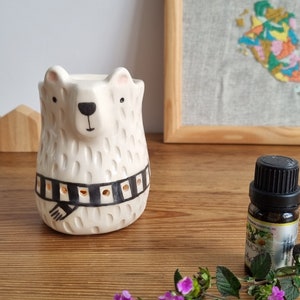 Puede incluir: Calentador de aceite de cerámica blanco con forma de oso con un diseño de bufanda a rayas blancas y negras. El calentador tiene un pequeño pozo para sostener una vela de té. También se puede ver una pequeña botella de aceite esencial en la imagen.