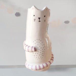 May include: A ceramic cat-shaped vase in a pale cream color. The cat features a hand-painted face with black eyes, nose, and whiskers. Its body is decorated with a central pattern of grey dots and pinkish-brown striped arms and a tail.