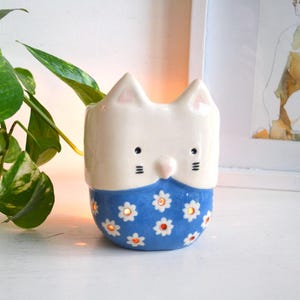 以下が含まれることがあります： 猫の形をした陶器製のキャンドルホルダー。上部は白く、尖った耳と描かれた顔が特徴です。下部は青色で、白いデイジーの模様と、光が漏れる小さな円形の切り抜きが施されています。