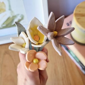 Puede incluir: Un pequeño ramo de flores de cerámica hechas a mano en varios colores. El ramo incluye flores blancas, amarillas, melocotón y marrones con centros amarillos. Las flores se sostienen en la mano de una persona, con una pintura y una vela en el fondo.