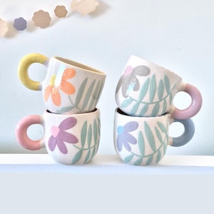 Puede incluir: Cuatro tazas de cerámica apiladas con diseños florales. Cada taza tiene un asa de diferente color: amarillo, rosa, gris y azul. Las tazas están decoradas con flores coloridas y hojas verdes.