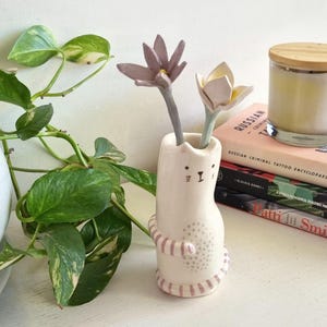 Puede incluir: Un jarrón de cerámica blanca con forma de gato y diseño de rayas rosas. Contiene dos flores artificiales, una lavanda y otra amarillo pálido. Una vela en un tarro de cristal y libros en el fondo. El jarrón está decorado con una cara de gato.