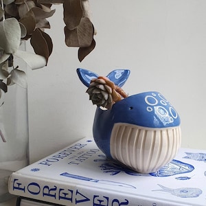 Puede incluir: Un macetero de cerámica azul y blanco en forma de ballena con una planta suculenta en su interior. La ballena tiene una barriga blanca con textura acanalada y una espalda azul con círculos blancos.