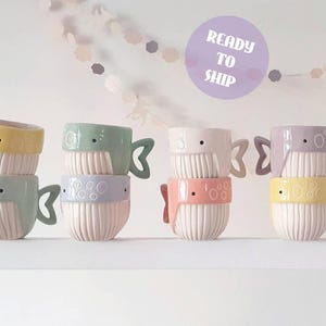 Puede incluir: Una colección de tazas de cerámica con forma de ballena en colores pastel, incluyendo amarillo, verde menta, lavanda y rosa. Cada taza presenta una base blanca acanalada, un asa pequeña con forma de cola de ballena y un ojo circular. Se observa un círculo morado con el texto READY TO SHIP.
