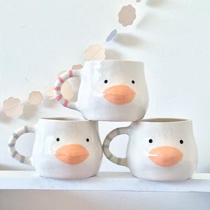 Puede incluir: Tres tazas de cerámica blanca apiladas sobre un estante blanco. Cada taza presenta un diseño de cara de pato con un pico naranja y ojos negros. Las asas están rayadas con colores pastel, incluyendo rosa, azul y verde. Una guirnalda decorativa cuelga en el fondo.