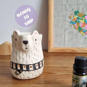 Puede incluir: Un quemador de aceite de cerámica blanca con forma de oso y superficie texturizada, que presenta un patrón de bufanda a rayas blancas y negras alrededor del centro. El oso tiene nariz y ojos negros, y una pequeña botella de aceite esencial cerca.