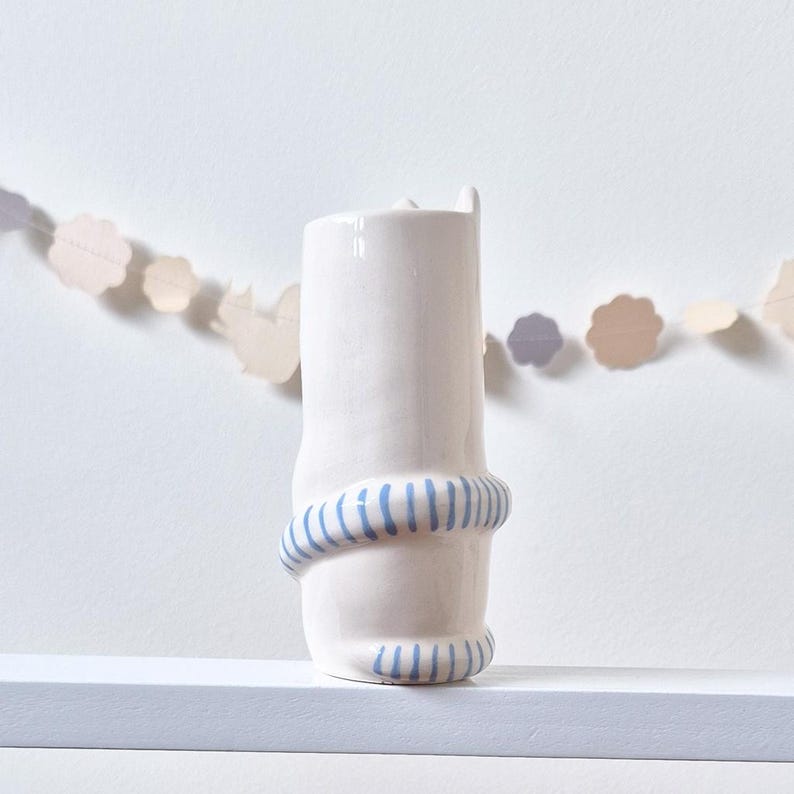 May include: A white ceramic vase with a glossy finish, featuring a sculpted tail wrapped around the lower section. The tail is decorated with horizontal light blue stripes. The vase has a cylindrical shape with a slightly irregular, organic texture.