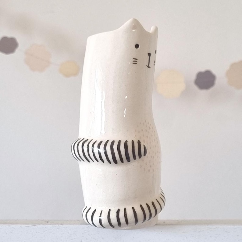 May include: A cream-colored ceramic vase shaped like a cat with a glossy finish. The cat features black painted details including eyes, whiskers, a nose, and striped patterns on its front paws and tail. A small dotted pattern is visible on the belly.