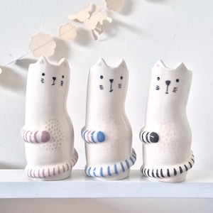 Puede incluir: Tres figuras de gato de cerámica blanca con detalles faciales negros y acentos a rayas. Cada gato tiene un diseño de rayas de diferentes colores alrededor de su centro y base, incluyendo rayas rosas, azules y negras. Se exhiben en una estantería blanca.