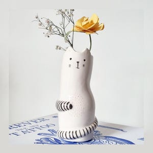 May include: A white ceramic cat-shaped vase with black hand-painted facial features, including eyes, whiskers, and a nose. The cat has black and white striped arms and a tail, with a textured dotted pattern on its belly, holding a yellow flower.