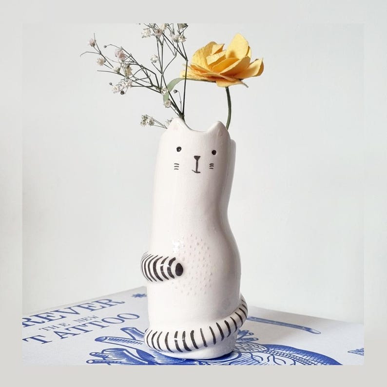 May include: A white ceramic cat-shaped vase with black hand-painted facial features, including eyes, whiskers, and a nose. The cat has black and white striped arms and a tail, with a textured dotted pattern on its belly, holding a yellow flower.
