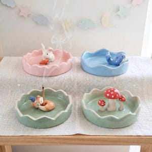 May include: Four pastel-coloured ceramic incense holders. Each features a different whimsical figurine: a bunny with a carrot, a whale, a fox, and mushrooms. Smoke rises from an incense stick in one holder.