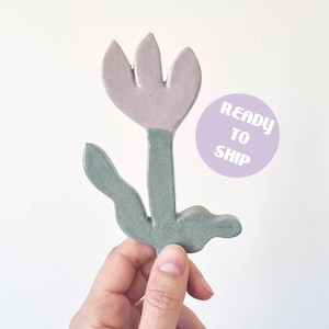 Puede incluir: Un adorno de cerámica en forma de flor con una flor de color malva claro de tres pétalos sobre un tallo verde salvia con dos hojas. Una pegatina circular de color violeta claro muestra el texto blanco READY TO SHIP en negrita.