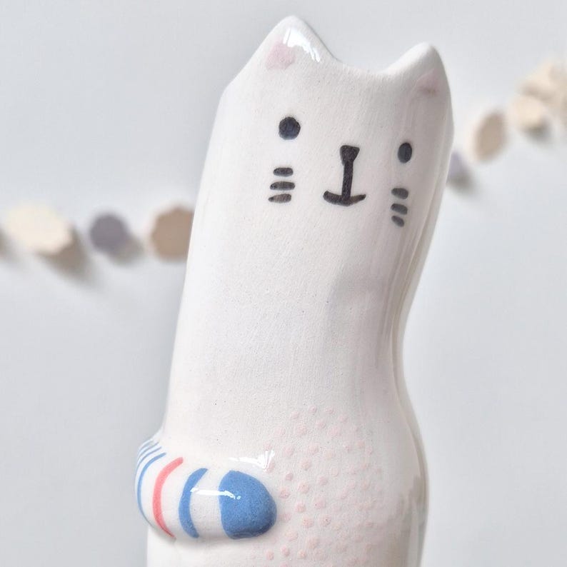 May include: A white ceramic cat figurine with a glossy finish. The cat has a hand-painted face with black eyes, a nose, and whiskers. A small tail wraps around the side, featuring horizontal stripes in light blue and coral pink colors.