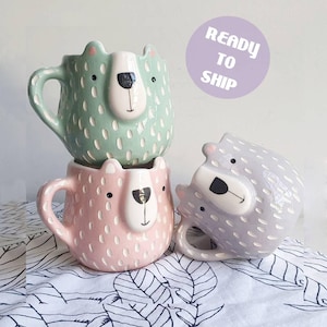Puede incluir: Tres tazas de cerámica con forma de caras de oso, una en verde menta, una en rosa claro y otra en lavanda. Cada taza presenta un hocico blanco, nariz negra y orejas pequeñas, con un patrón texturizado de rayas blancas en la superficie.