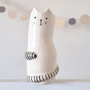 May include: A white ceramic cat-shaped vase with a glossy finish. The cat features a hand-painted black face with simple eyes, a nose, and whiskers. It has a striped black and white tail wrapping around the base and a striped paw on its side.