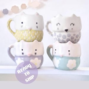 Puede incluir: Cuatro tazas de cerámica con forma de animal y orejas. La taza superior izquierda es amarilla con rayas blancas, la superior derecha es gris con puntos, la inferior izquierda es lavanda con flores blancas y la inferior derecha es azul claro con flores blancas.