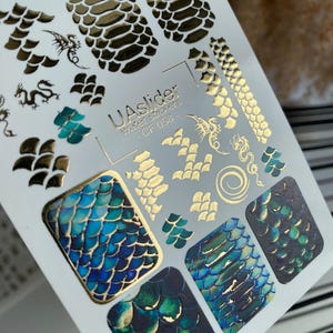 Peut inclure: Une planche d'autocollants pour ongles à transfert d'eau. Les autocollants présentent des motifs d'écailles de dragon dorées et turquoise, ainsi que des motifs de dragons et d'écailles. Le texte "UAslider water stickers - CF 058" est imprimé.