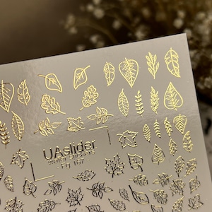 May include: Sheet of nail art water stickers featuring various gold leaf designs. The sheet includes different leaf shapes and the text "UASlider water stickers - Fg 167". The stickers are on a clear background.