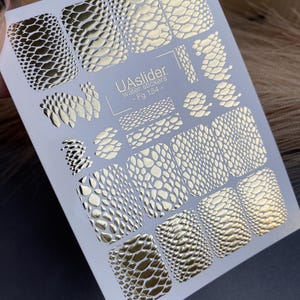 Könnte beinhalten: Ein Bogen mit Wassertransfer-Nagelaufklebern mit verschiedenen goldenen Reptilienhautmustern. Der Bogen enthält den Text "UASlider water stickers - Fg 184 -."