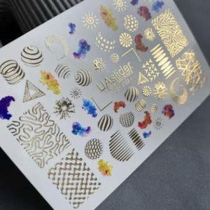 May include: A sheet of nail art stickers with various gold foil designs, including suns, moons, and geometric shapes. The stickers also feature colorful watercolor-like accents. The text "UA slider" is visible on the sheet.