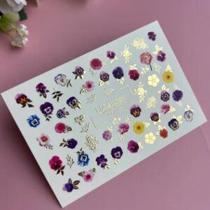 Pode incluir: Uma folha de autocolantes para unhas com uma variedade de desenhos de flores de amor-perfeito coloridas em tons de roxo, rosa, amarelo e azul, com detalhes em folha de ouro. Os autocolantes estão dispostos sobre um fundo branco.