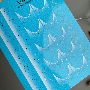 Könnte beinhalten: Ein Bogen blauer Wasseraufkleber mit einem Wellenmuster. Der Text "UASLIDER water stickers - CF 768" ist oben aufgedruckt. Die Aufkleber zeigen weiße Wellenmuster und kleine, verstreute Punkte.