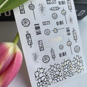 May include: A sheet of nail art stickers featuring black and gold geometric designs. The designs include circles, rectangles, and linear patterns with a textured, mosaic-like appearance. The text "UAslider Winter Stickers - OF 476-" is printed on the sheet.