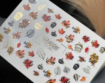 Puede incluir: Hoja de pegatinas para uñas con diseños de temática otoñal. Incluye hojas de otoño rojas, naranjas y doradas, tazas de café y la palabra "LOVE". También está presente el texto "Hello Fall".
